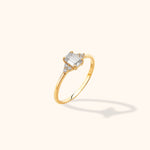 Gold-colored ring with a rectangular clear center stone, small round side stones, and a slim polished band on a cream background.