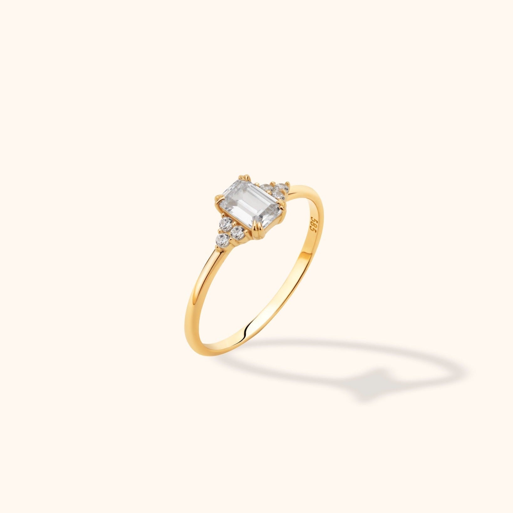 Gold-colored ring with a rectangular clear center stone, small round side stones, and a slim polished band on a cream background.