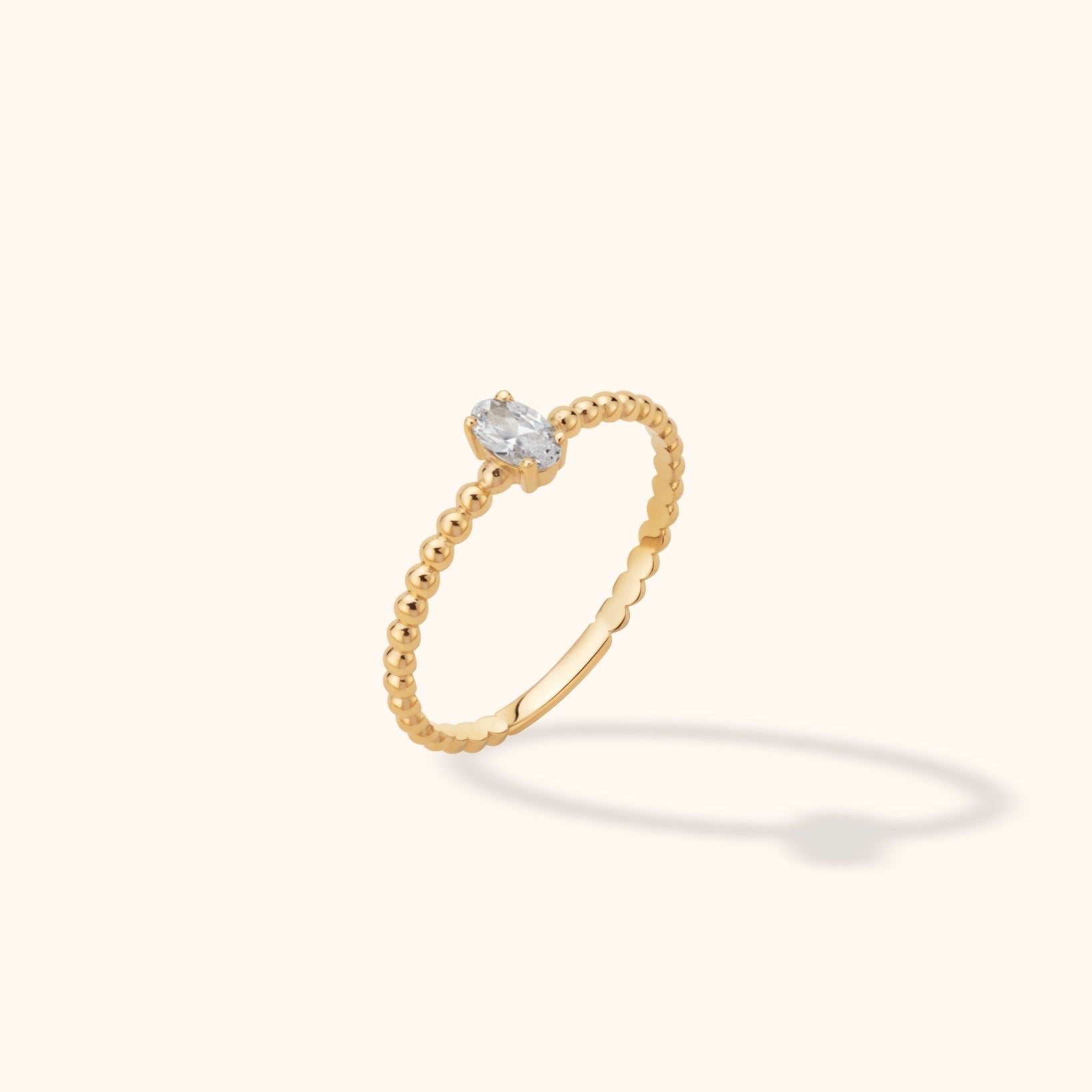 Gold-toned ring with a beaded band and a clear oval stone in a prong setting, shown at an angle on a cream background.