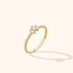 Gold-colored ring with a slim beaded band and three clear rectangular stones in an angled cluster, shown on a cream background.