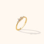 Slim gold-colored ring with staggered round clear stones in varied sizes, shown at an angle on a pale cream background.