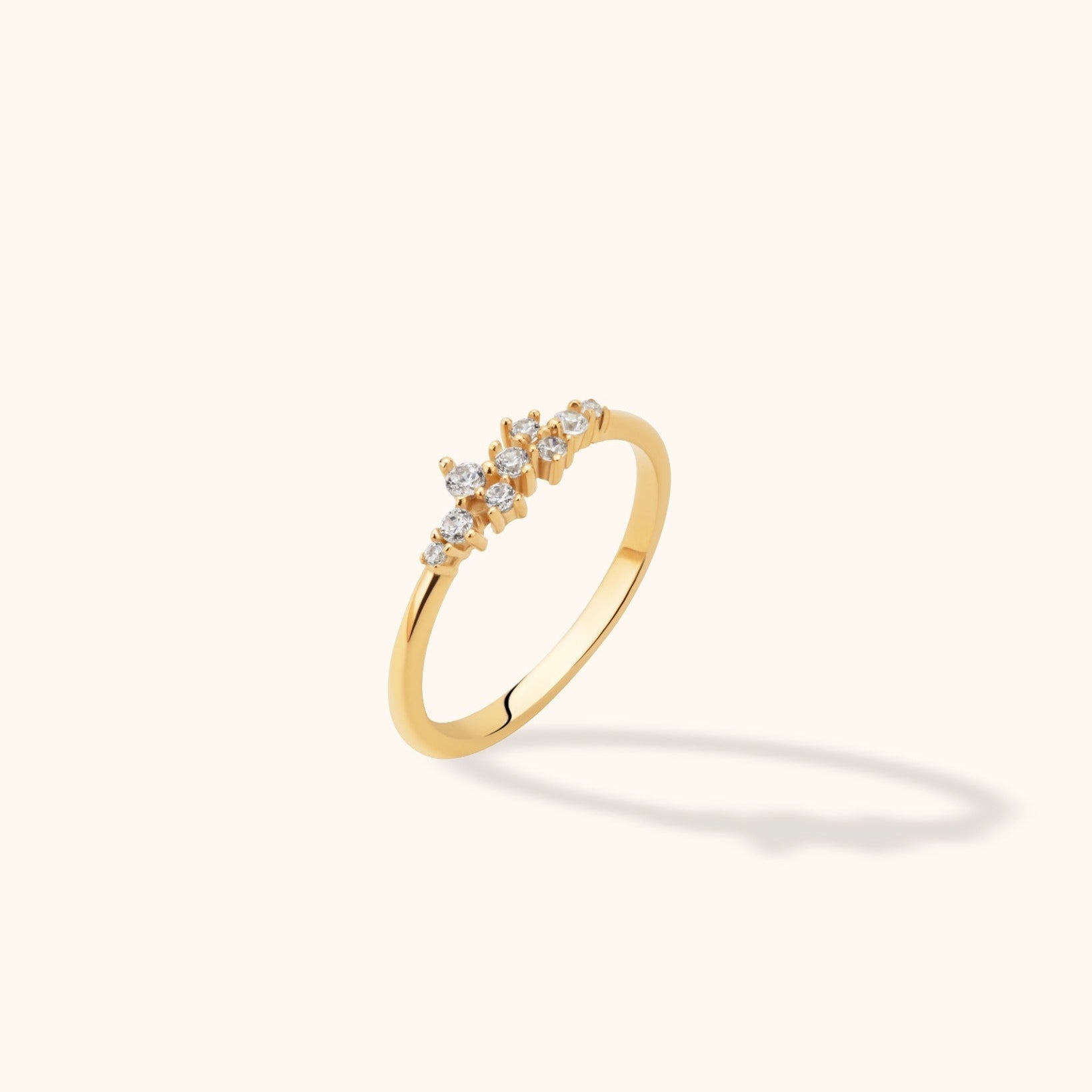 Slim gold-colored ring with staggered round clear stones in varied sizes, shown at an angle on a pale cream background.