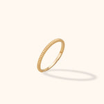 Slim gold-colored ring with a ribbed outer edge and smooth inner surface, tilted against a cream background with a soft shadow.