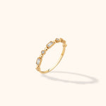 Slim gold-colored ring with round and rectangular clear stones, polished bead accents and ridged settings on a cream background.
