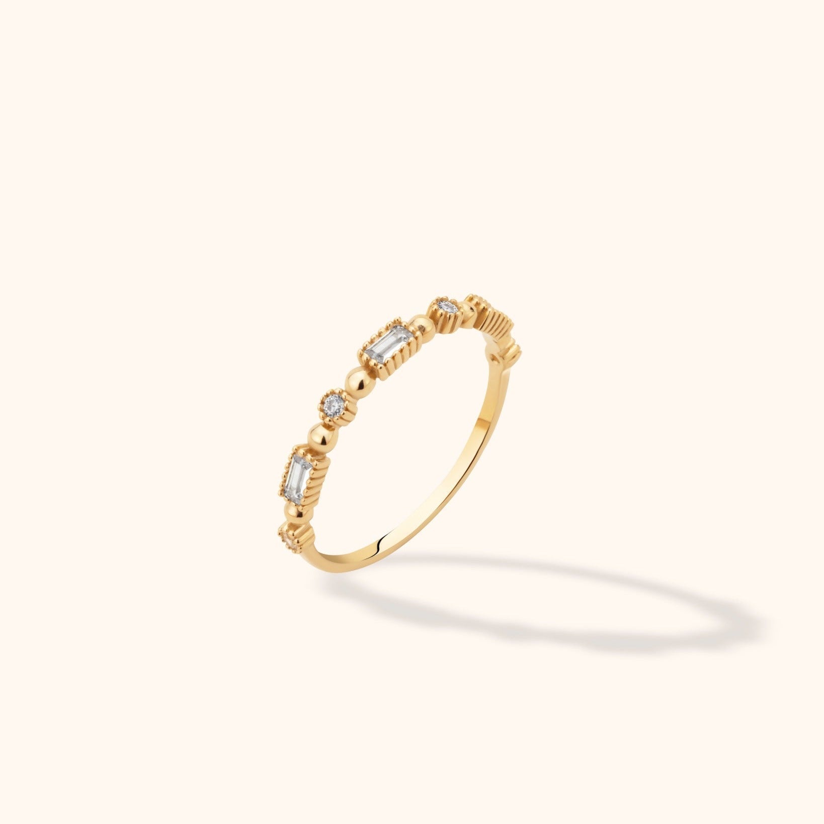 Slim gold-colored ring with round and rectangular clear stones, polished bead accents and ridged settings on a cream background.