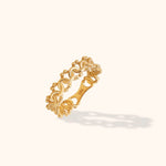 Gold-colored ring with interwoven loops and small rounded edge details, shown tilted on a cream background.
