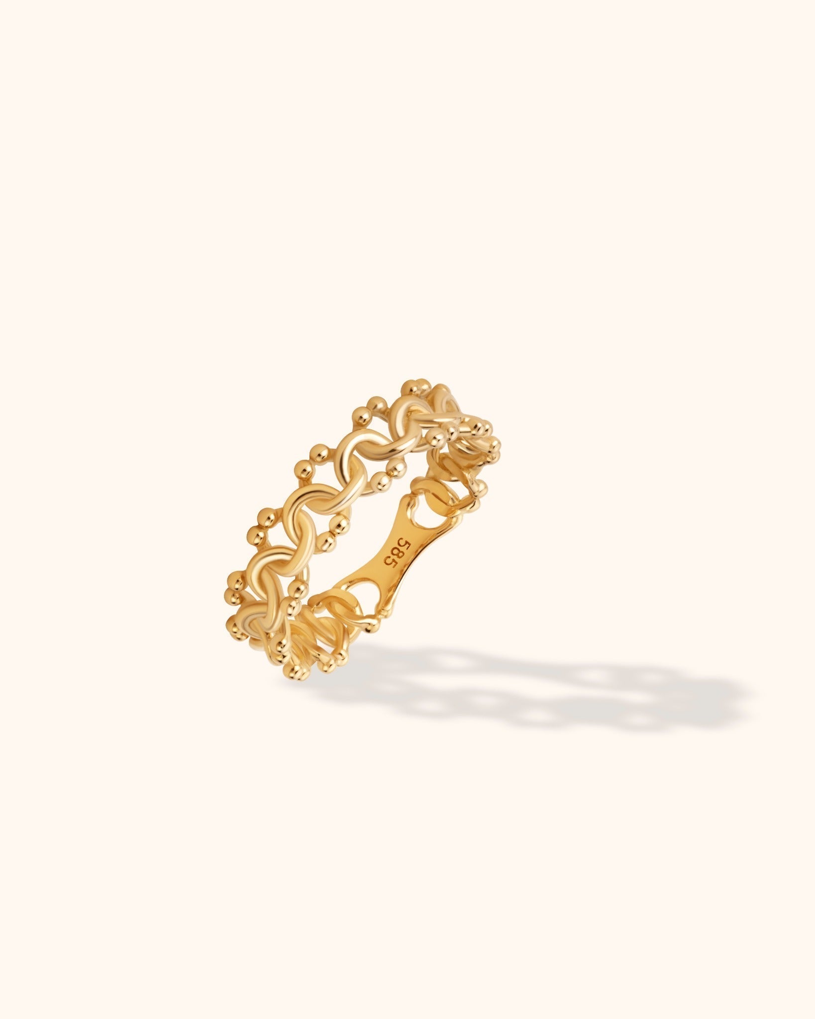 Gold-colored ring with interwoven loops and small rounded edge details, shown tilted on a cream background.