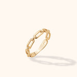 Gold-toned ring with elongated paperclip-style links and a smooth lower band, tilted against a cream background.