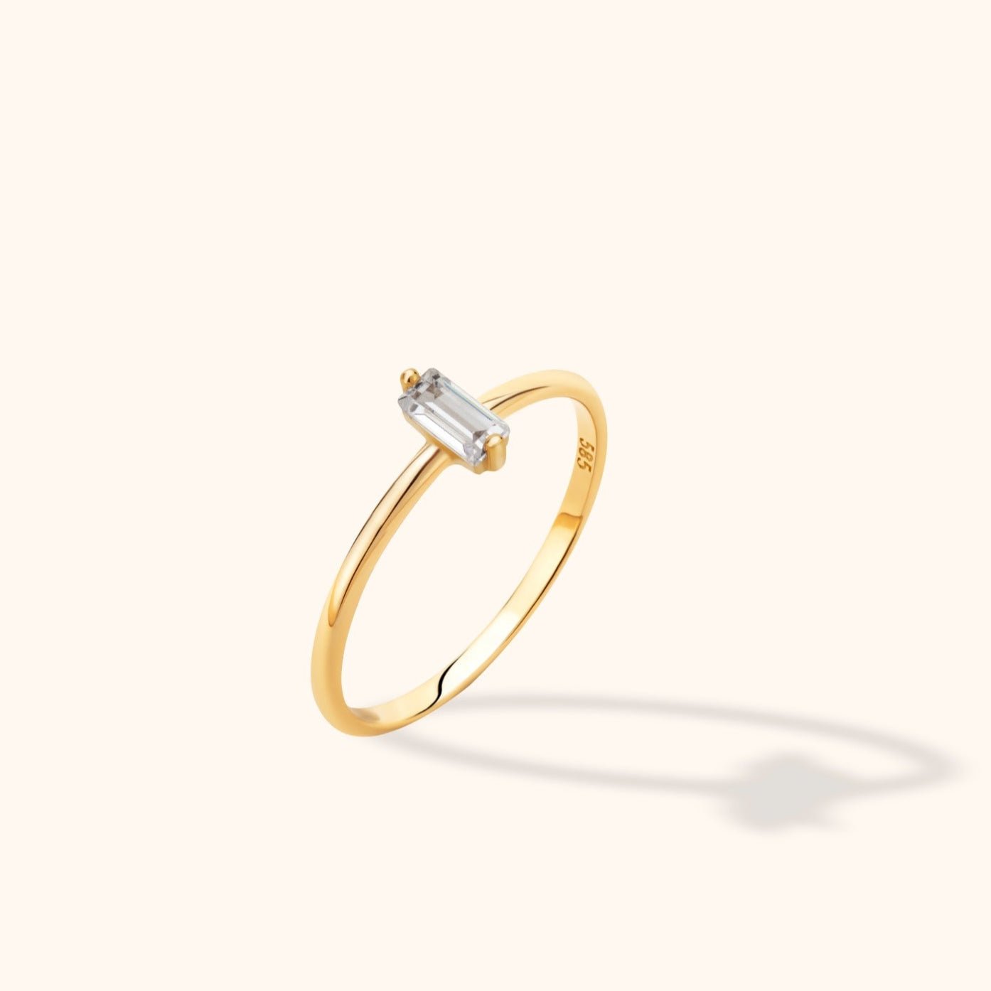 Slim gold-colored ring with a clear rectangular stone in a prong setting, shown at an angle on a pale cream background.