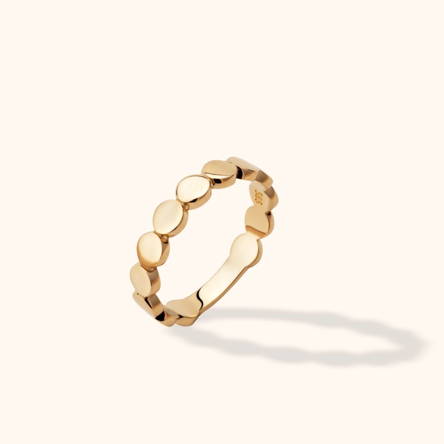 Shiny gold-toned ring with rounded pebble-like segments, shown tilted upright on a cream background with a soft shadow.