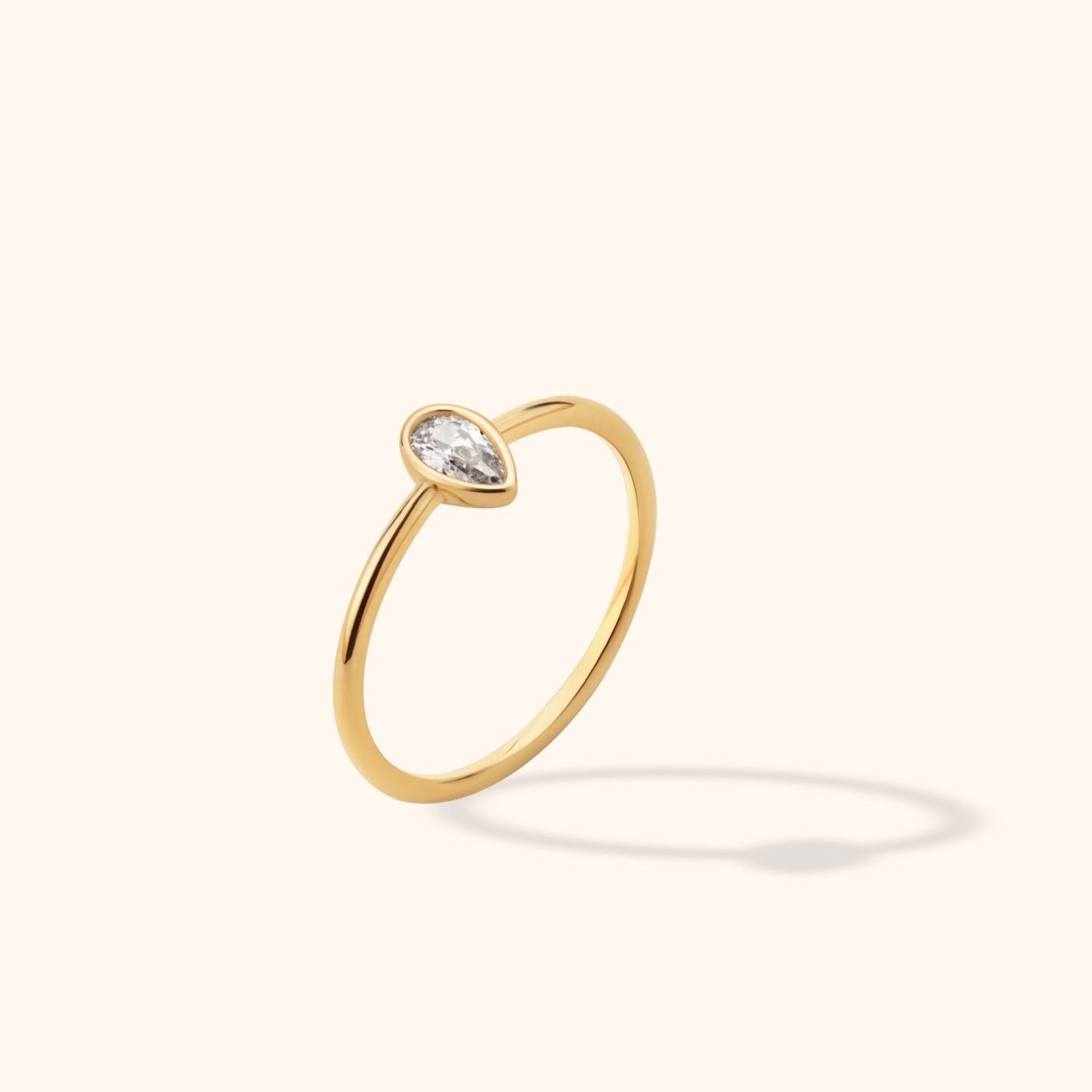 Slim gold-toned ring with a clear pear-shaped stone in a smooth bezel setting, shown at an angle on a cream background.
