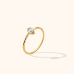 Slim gold-toned ring with a clear pear-shaped stone in a smooth bezel setting, shown at an angle on a cream background.