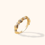 Gold-colored ring with rounded ribbed sections and small clear accents, shown tilted on a cream background with a soft shadow.