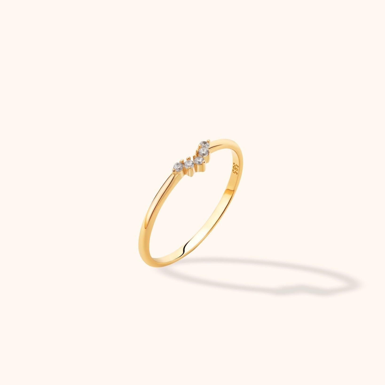 Slim gold-colored ring with five round clear stones along a shallow V-shaped front, shown at an angle on a cream background.