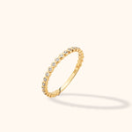Slim gold-toned ring with a row of clear round accents in circular settings, shown tilted against a pale cream background.