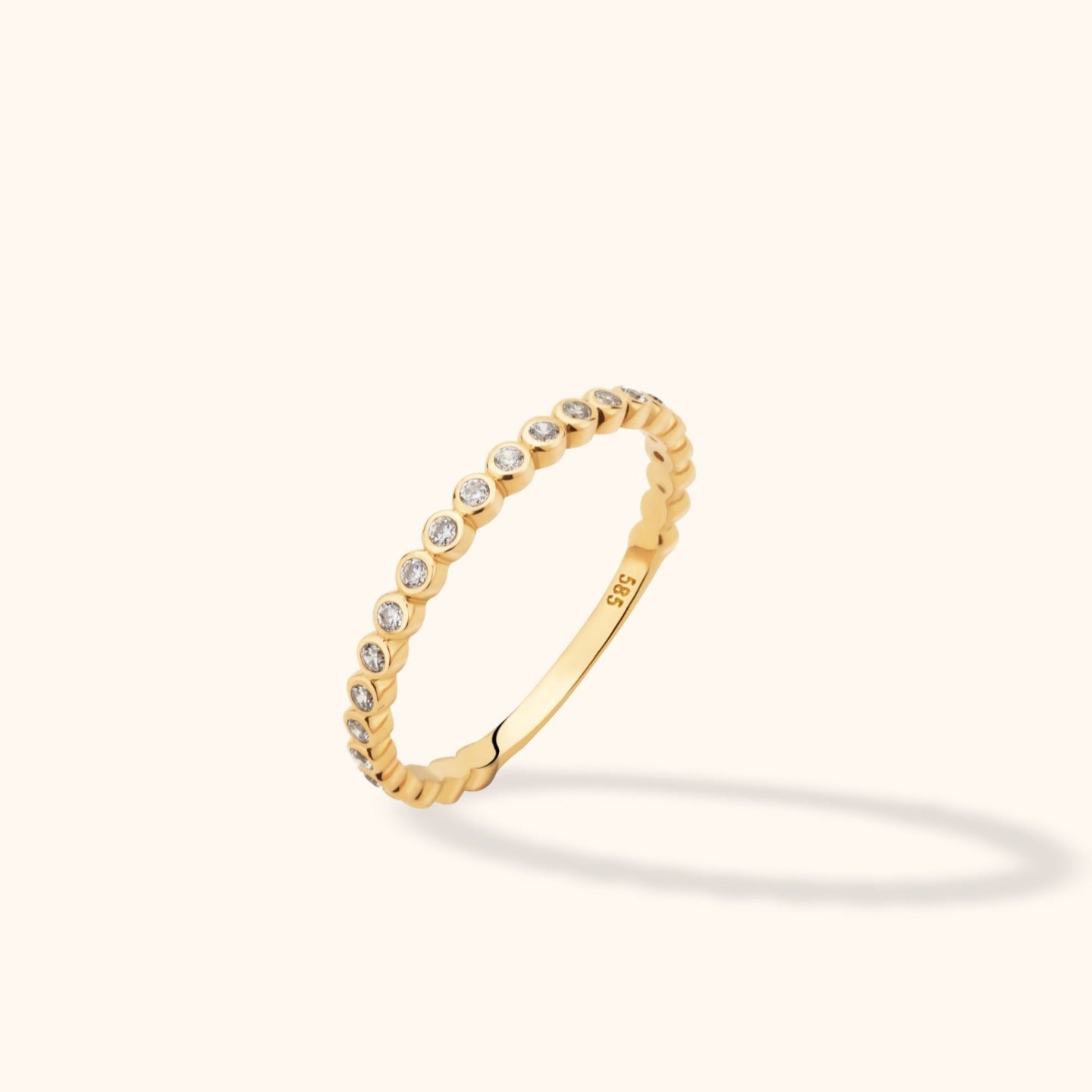 Slim gold-toned ring with a row of clear round accents in circular settings, shown tilted against a pale cream background.