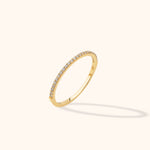 Slim gold-toned ring with a single row of small clear stones along the upper arc, tilted against a cream background.