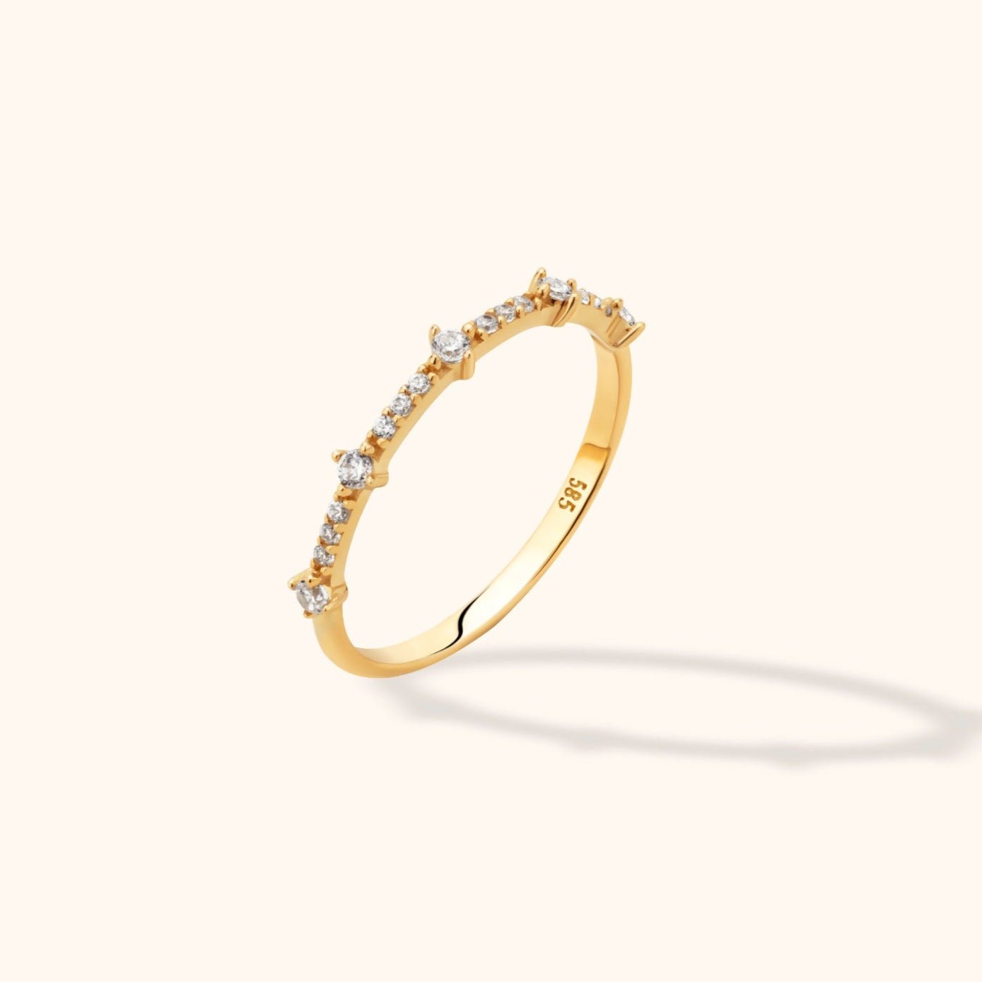 Slim gold-colored ring with larger round clear accents spaced between rows of smaller accents, shown tilted on a cream background.