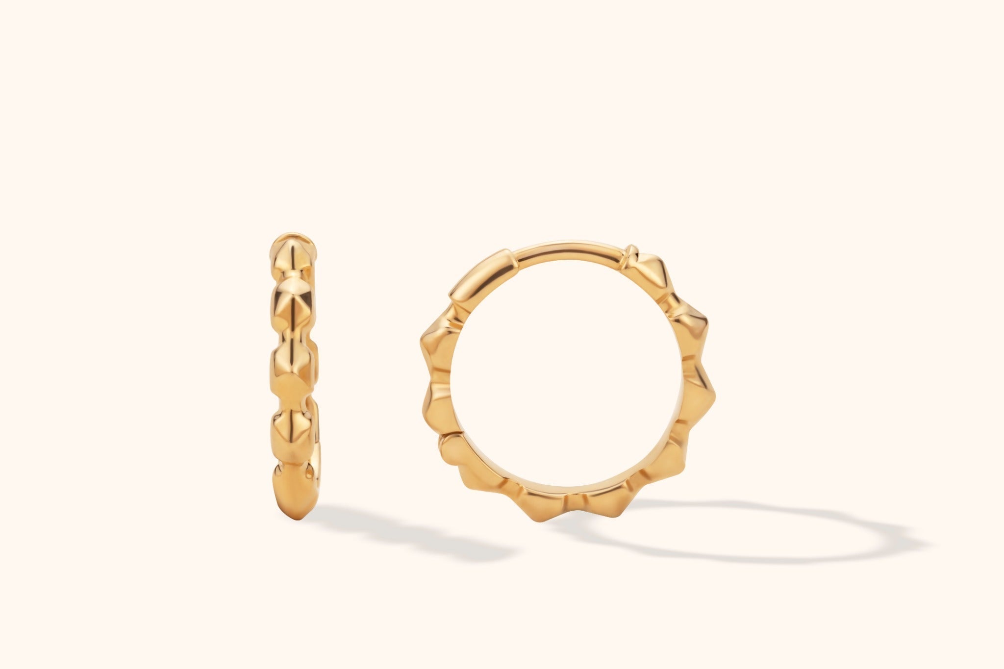 Two polished gold-tone hoop earrings with angular, sculpted edges, shown face-on and edge-on against a cream background.