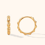 Two polished gold-tone hoop earrings with angular, sculpted edges, shown face-on and edge-on against a cream background.