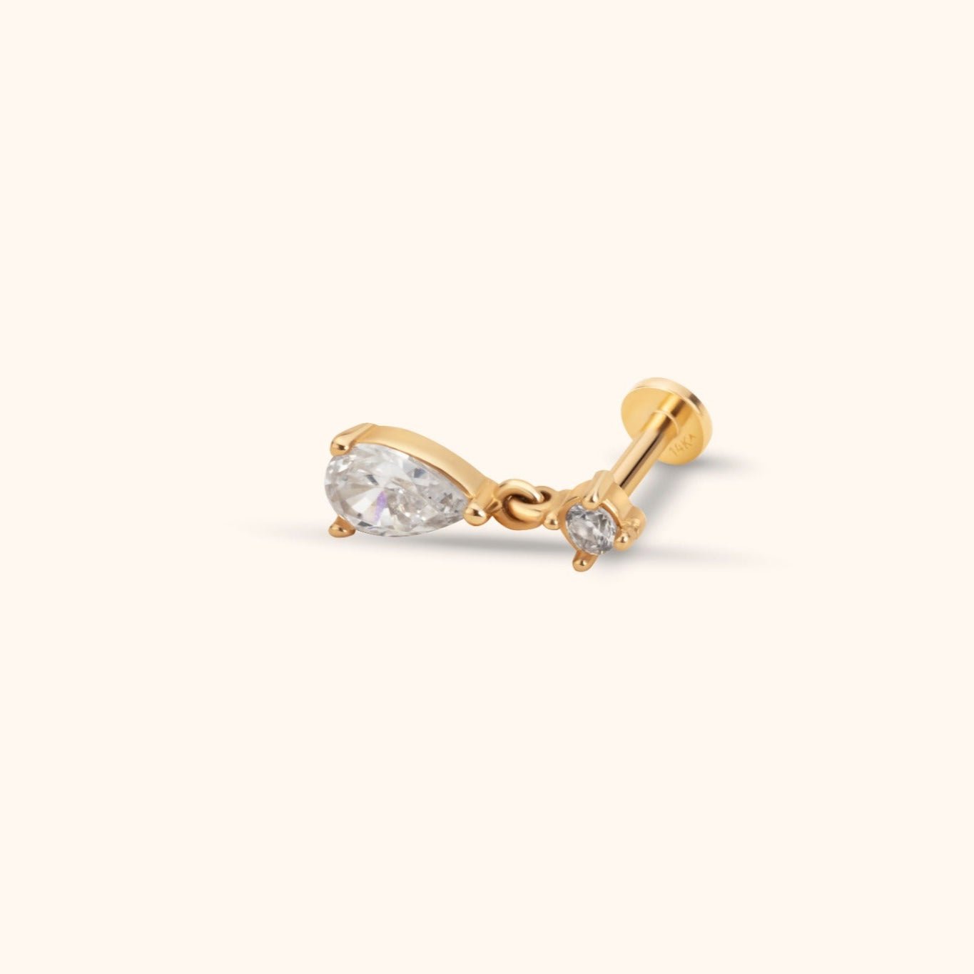 Gold-toned flat-back stud with a small round clear stone and a dangling pear-shaped clear stone on a cream background.