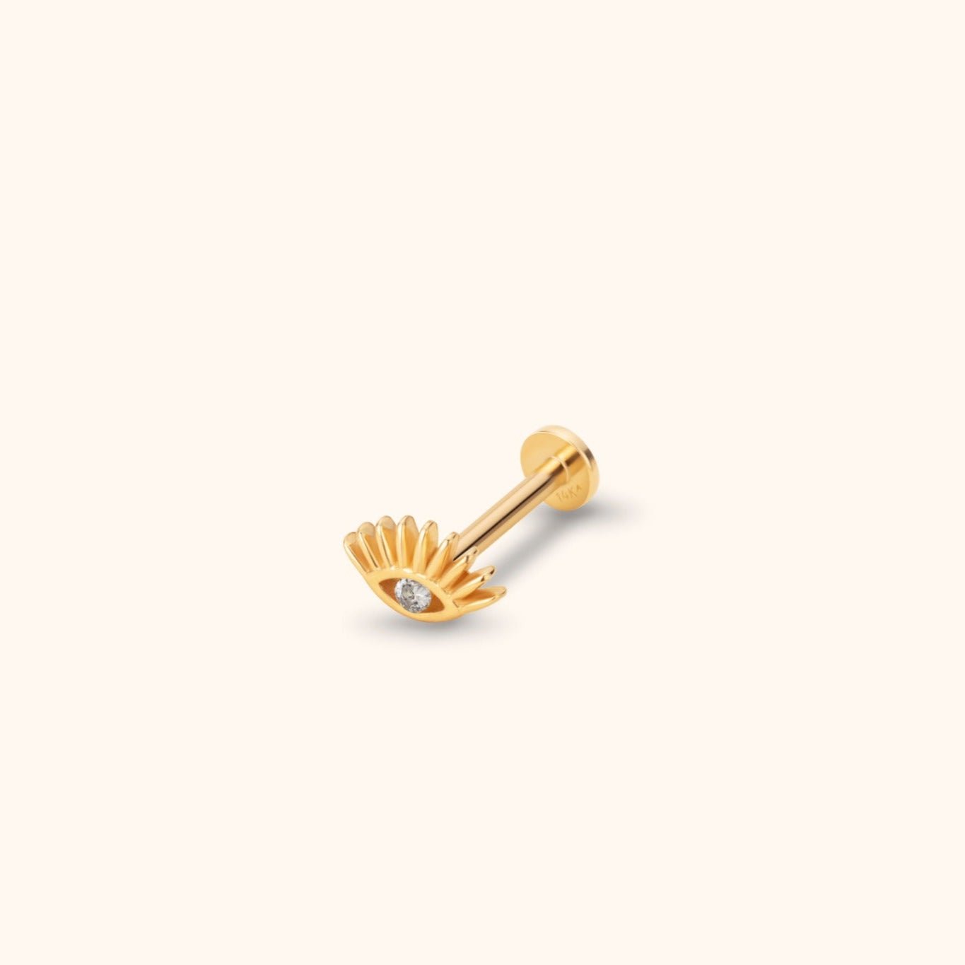 Gold-colored eye-shaped stud with radiating lashes, a clear faceted center, straight post and round flat back on a cream background.