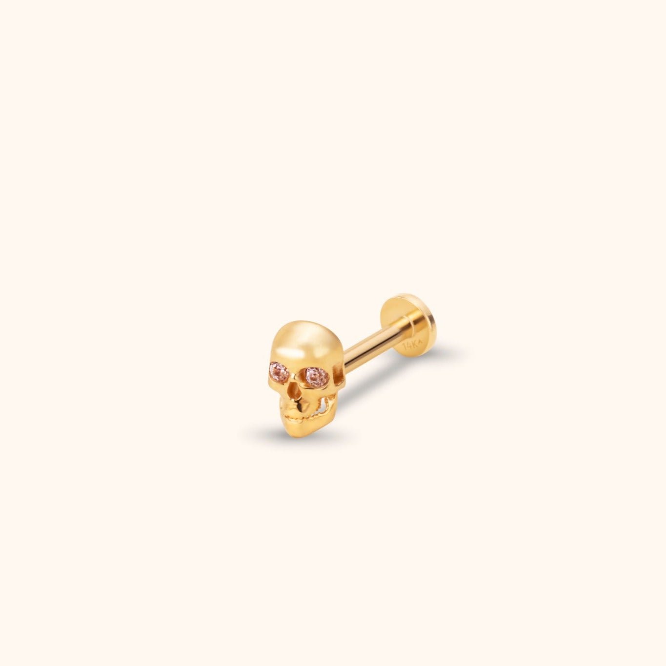 Gold-colored skull stud with pink faceted eyes, a straight post and a flat round back, shown at an angle on a cream background.