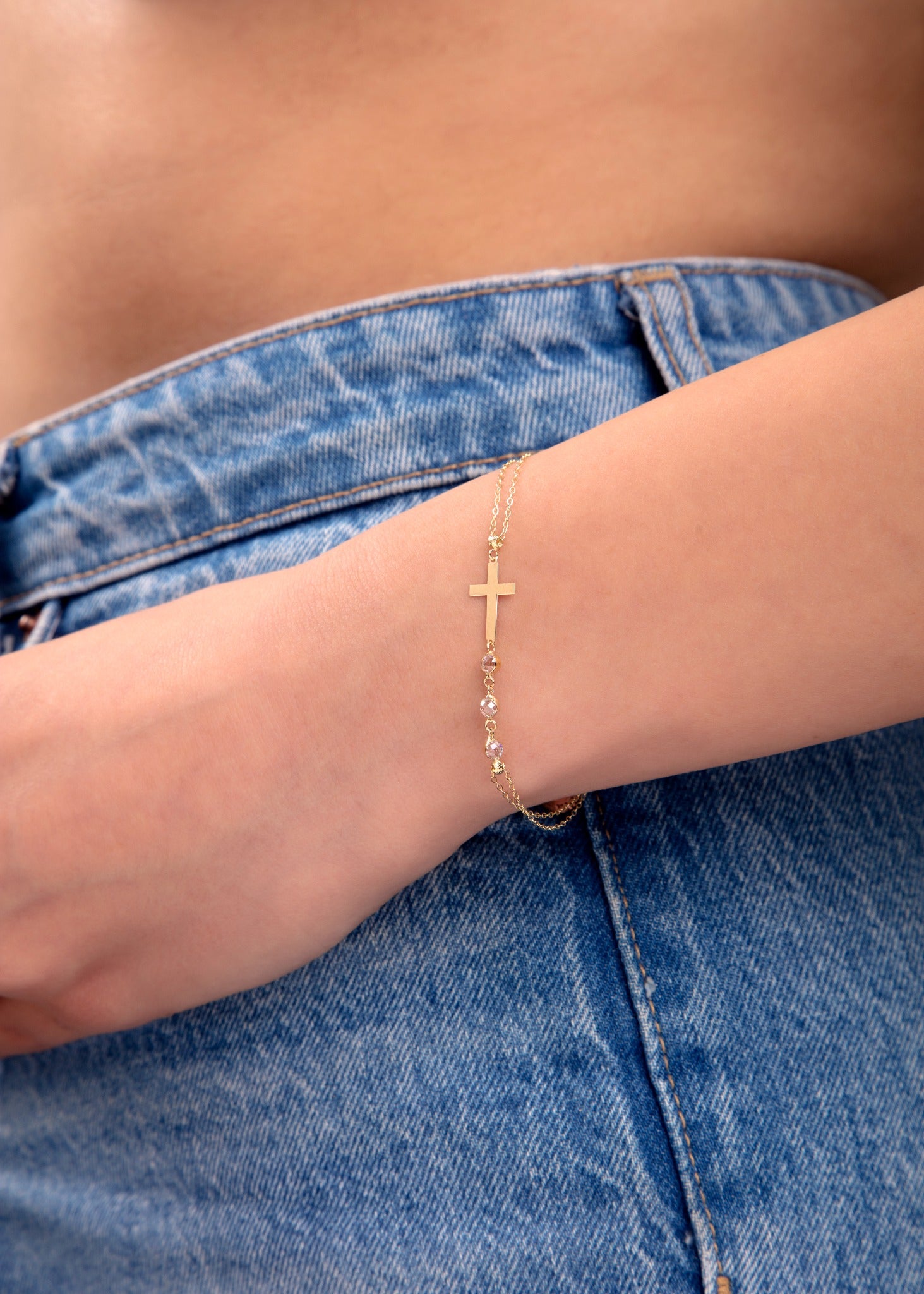 Minimal gold bracelet with cross charm and crystal beads on wrist, styled with denim

