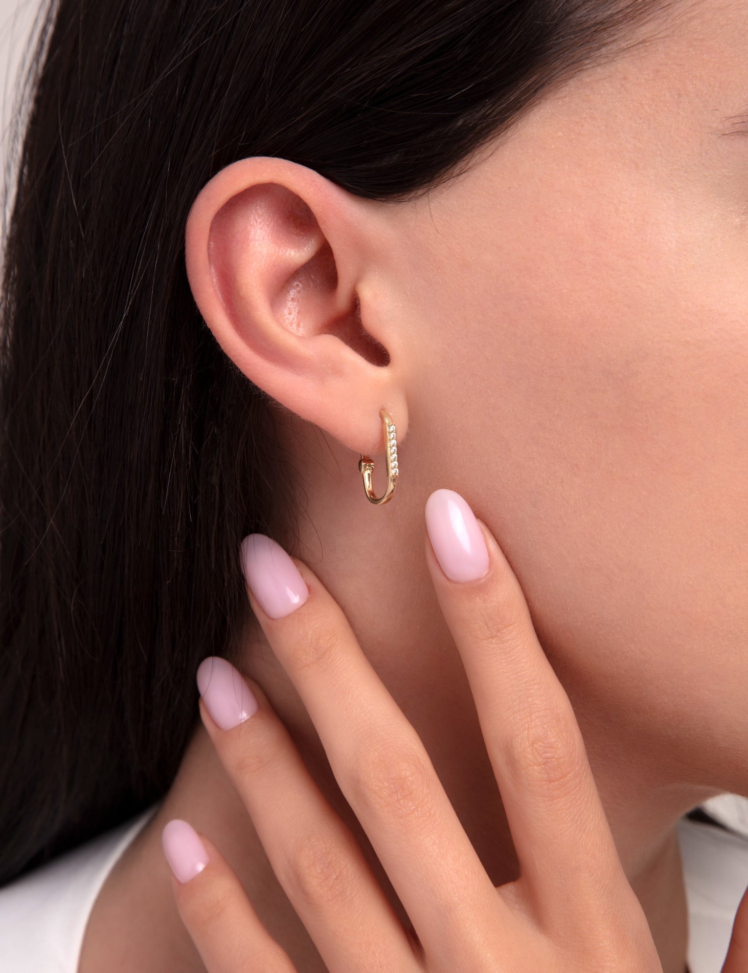 Close-up of a woman's ear wearing a 14K gold hoop earring with pavé zirconia stones, paired with polished pink nails.