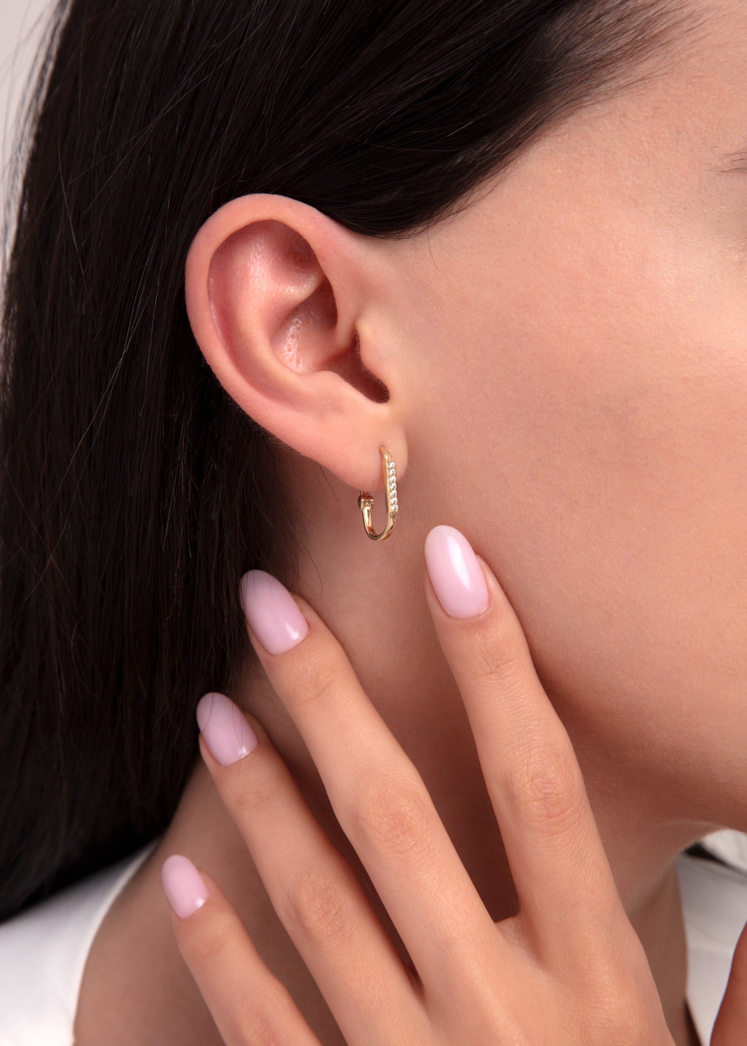 Close-up of a woman's ear wearing a 14K gold hoop earring with pavé zirconia stones, paired with polished pink nails.