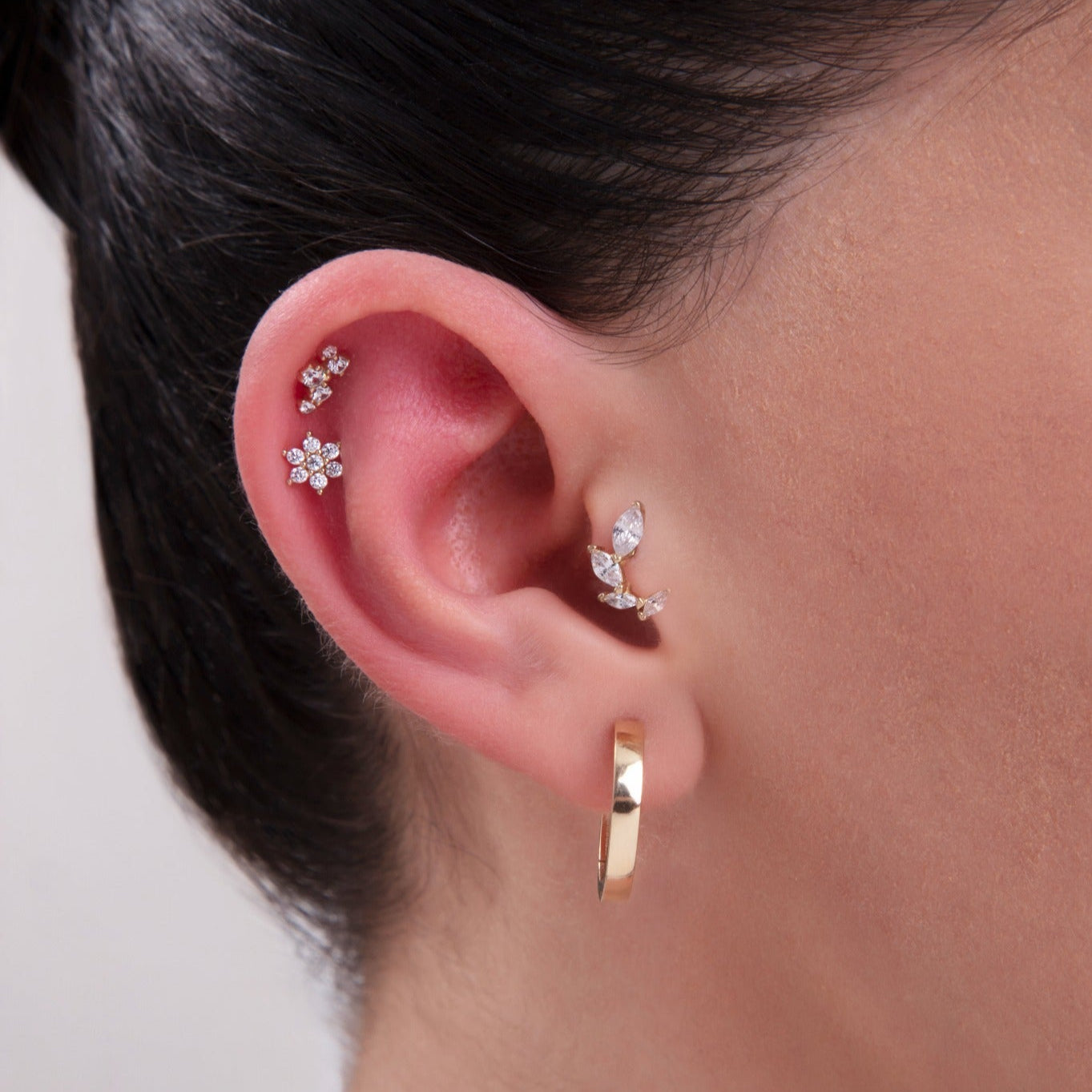Curated ear stack featuring a trio of 14K gold cartilage studs: a floral CZ cluster in the helix, a larger flower stud in the middle cartilage, and a marquise leaf-shaped CZ earring in the inner conch – Finished with a polished gold hoop in the lobe
