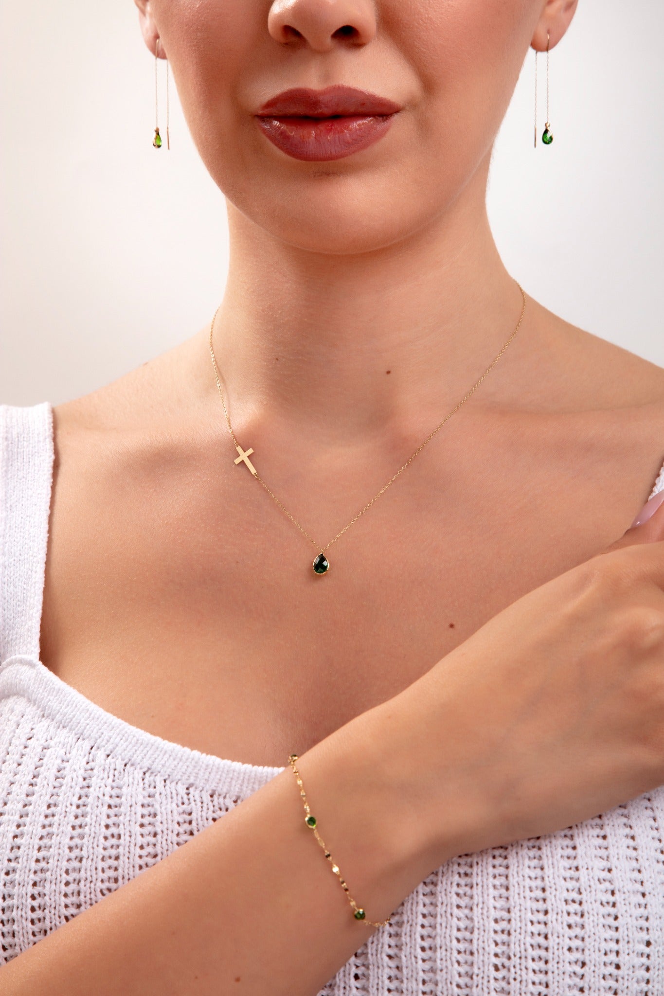 Woman wearing a 3-piece emerald green jewelry set including side cross necklace with teardrop, matching threader earrings, and dotted green stone bracelet

