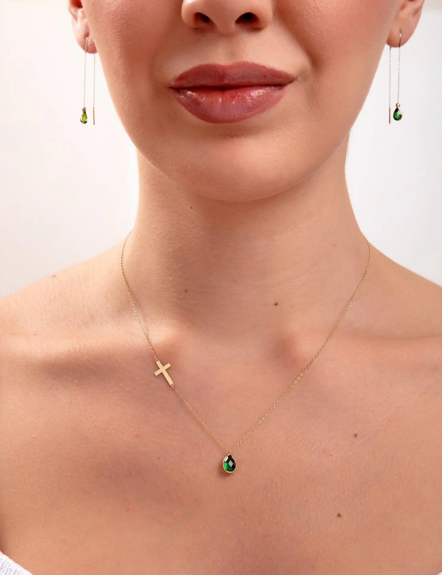Model wearing a 14K gold plated necklace with emerald teardrop crystal and horizontal gold cross, paired with matching green teardrop threader earrings

