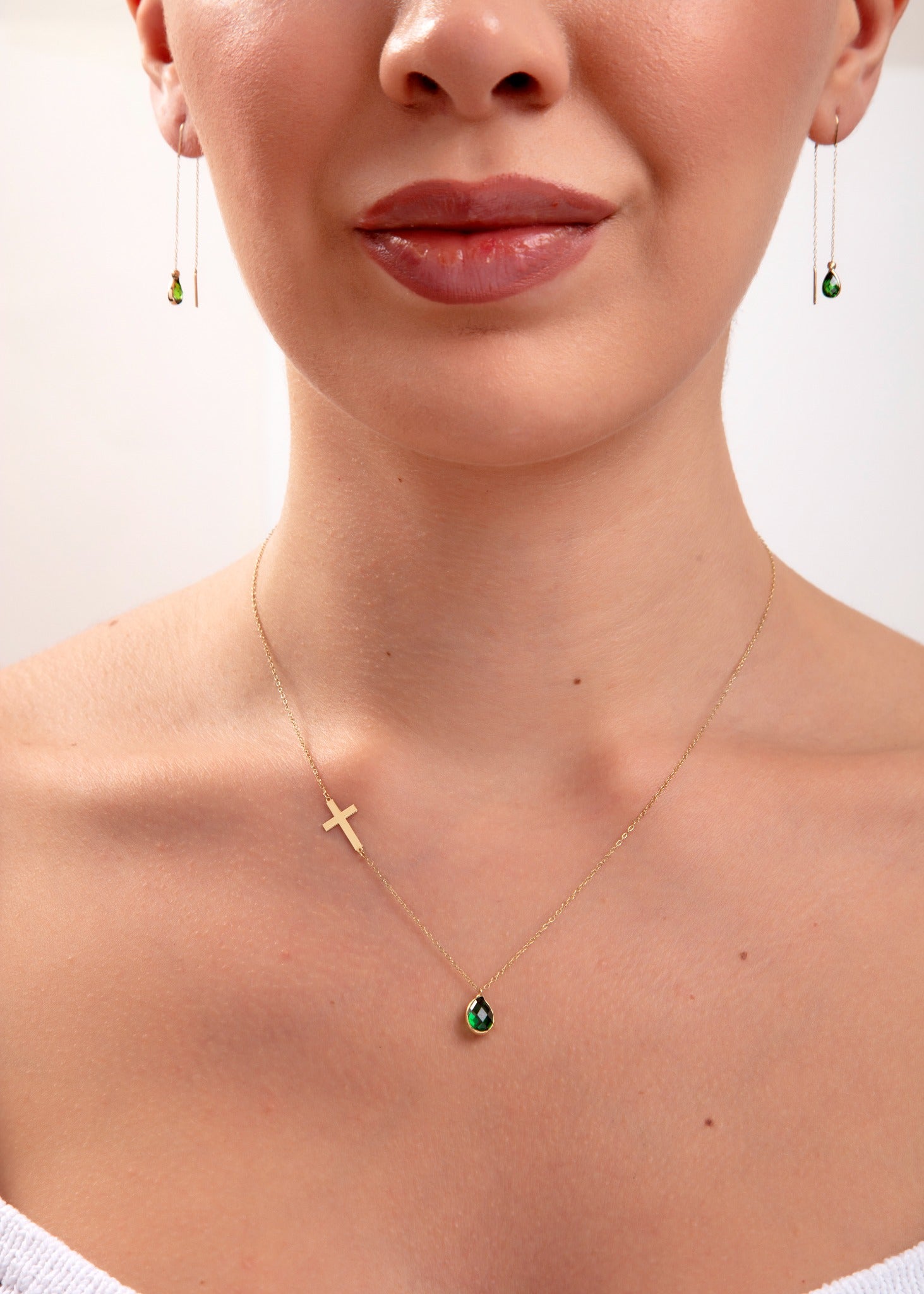 Model wearing a 14K gold plated necklace with emerald teardrop crystal and horizontal gold cross, paired with matching green teardrop threader earrings

