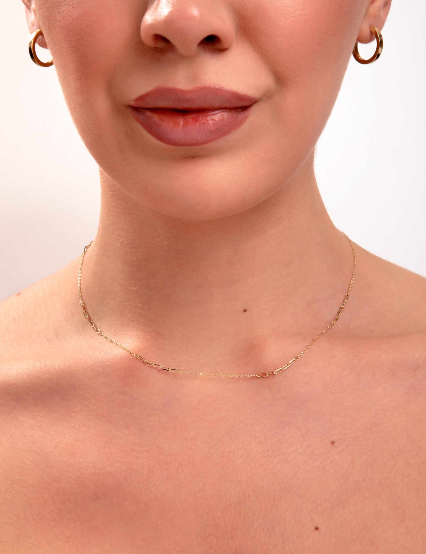 Close-up of minimalist gold paperclip chain necklace on model's neck, paired with small gold hoop earrings


