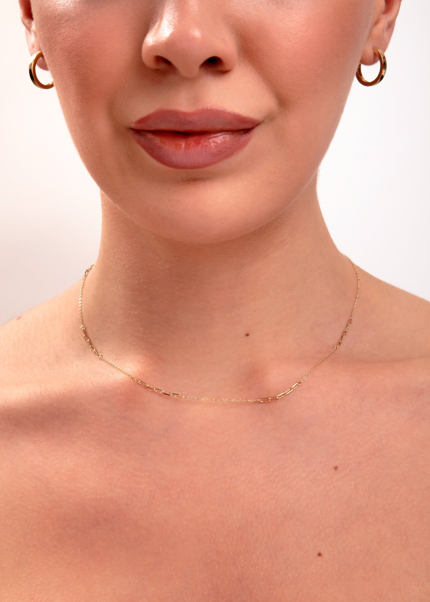 Close-up of minimalist gold paperclip chain necklace on model's neck, paired with small gold hoop earrings

