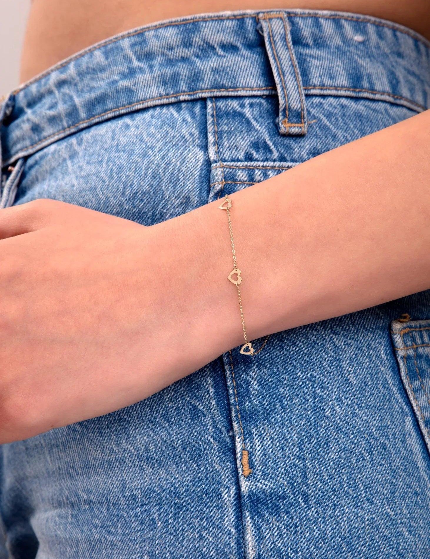 Delicate gold bracelet with open heart shapes, styled on wrist, paired with high-waisted blue jeans

