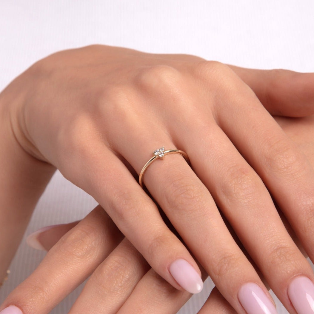 Delicate gold solitaire ring with a single sparkling stone on a woman's finger, styled with light pink manicured nails and gold bracelets, conveying elegance and simplicity.

