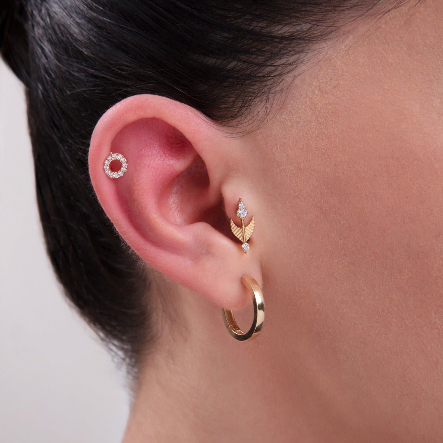 Model showcasing a curated ear stack featuring a 14K Gold Pavé CZ Circle Stud in the helix, an arrow-shaped CZ cartilage stud in the conch area, and a classic gold hoop in the lobe – A stylish layered piercing combination for modern ear styling