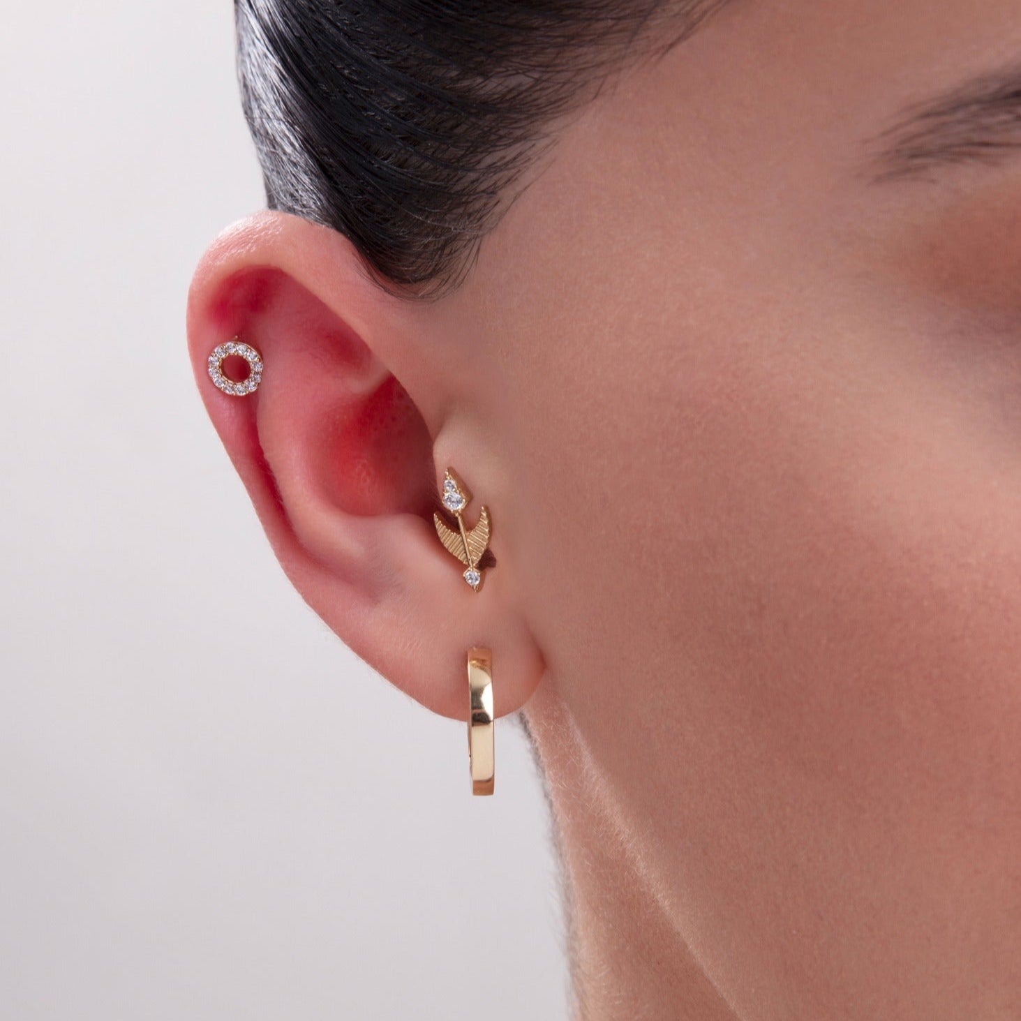 Model showcasing a layered ear stack with a 14K Gold Pavé Circle Helix Stud, a gold arrow-shaped cartilage earring with CZ accents, and a polished gold hoop in the lobe – A chic and symbolic curated ear combination