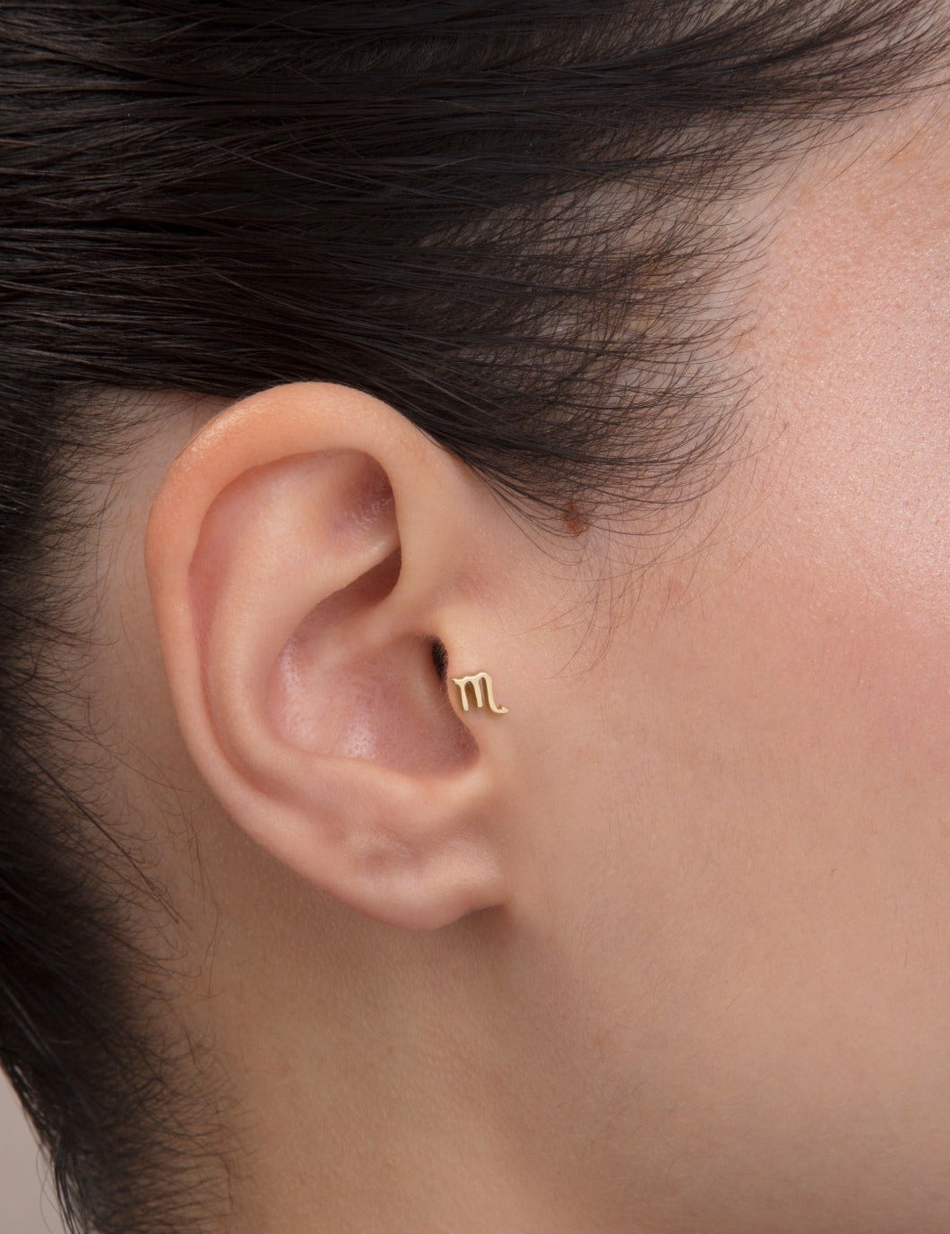 Yakın plan kulak; iç conch’ta “m” harfi formunda akrep mini altın burç küpesi.