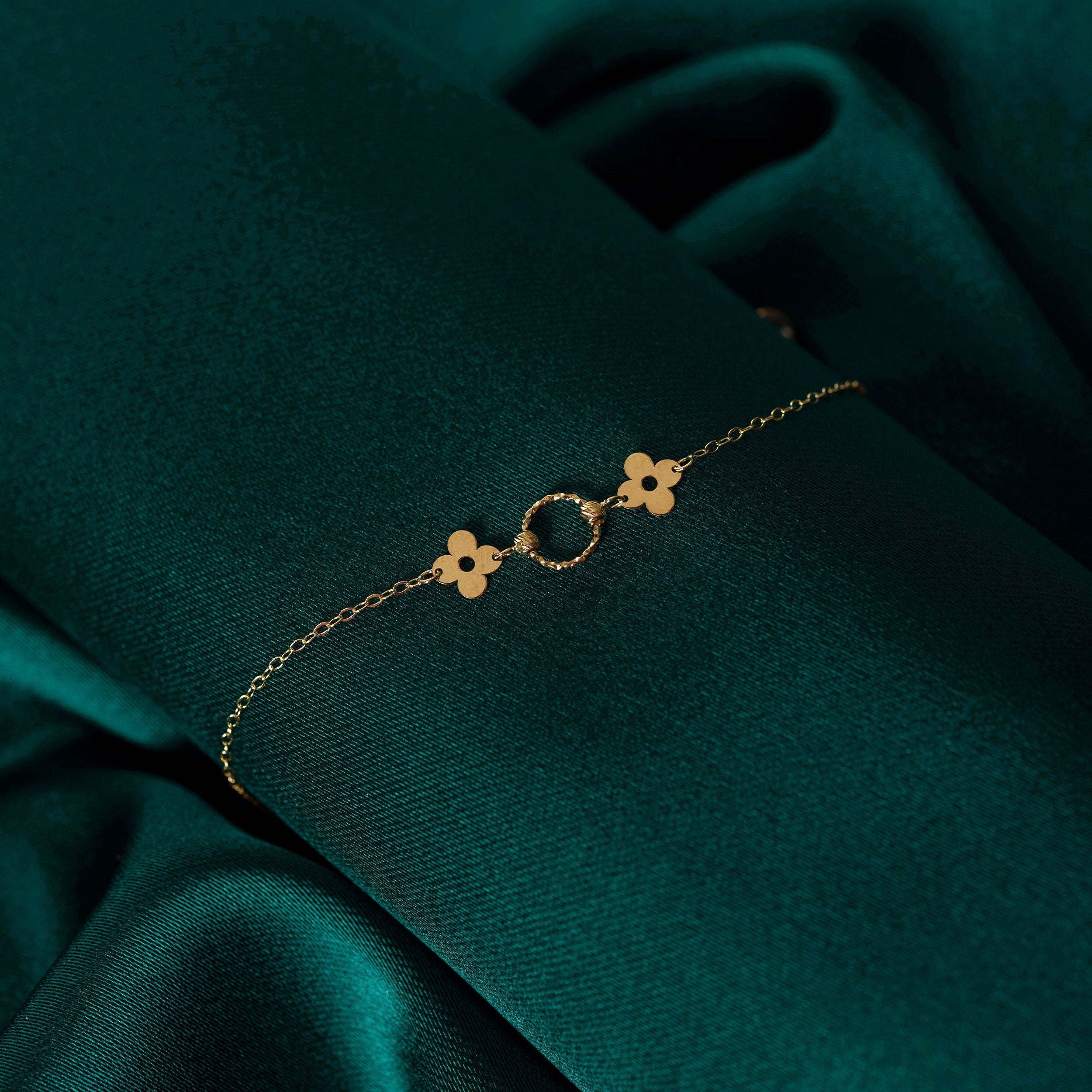 14K gold bracelet with a twisted circle centerpiece and two solid clover charms on a fine chain, displayed on deep green satin fabric.