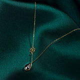 Elegant 14K gold necklace with black onyx teardrop pendant and floral charm on dark green fabric background