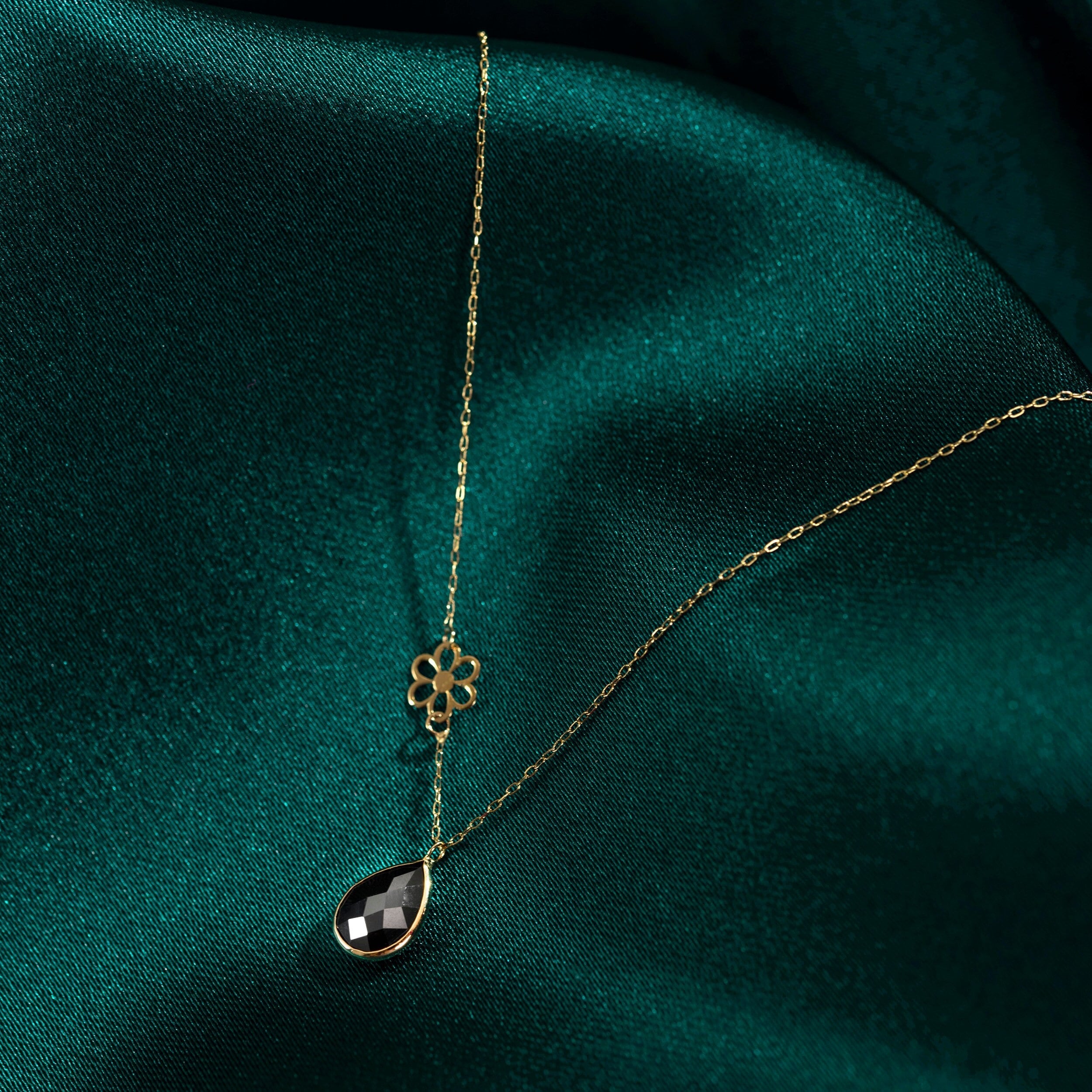 Elegant 14K gold necklace with black onyx teardrop pendant and floral charm on dark green fabric background