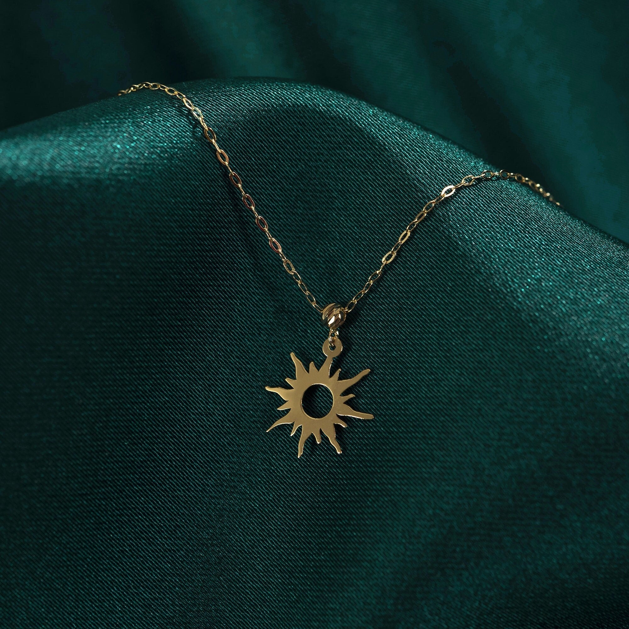 14K gold necklace featuring a radiant sunburst charm with pointed rays and a hollow center, styled on rich emerald green satin.