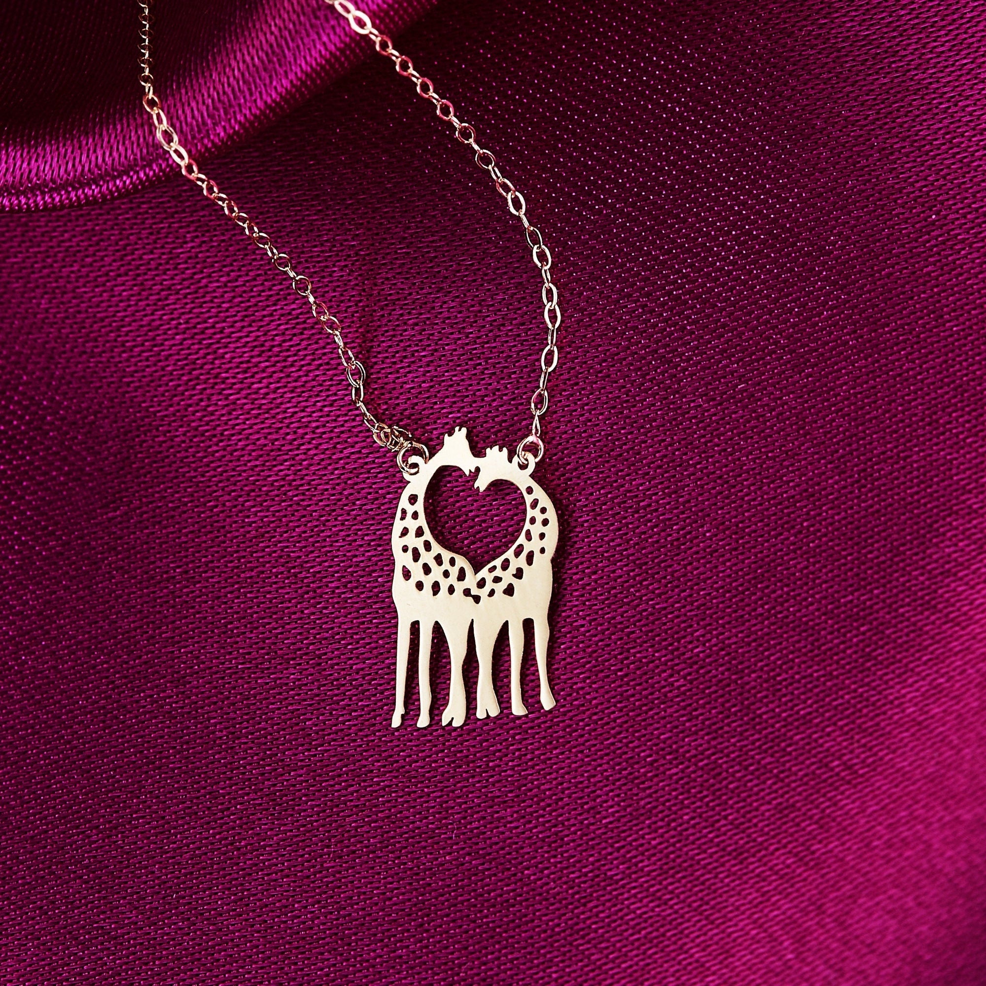 14K gold necklace with two giraffes forming a heart shape, laid on a magenta silk fabric background.