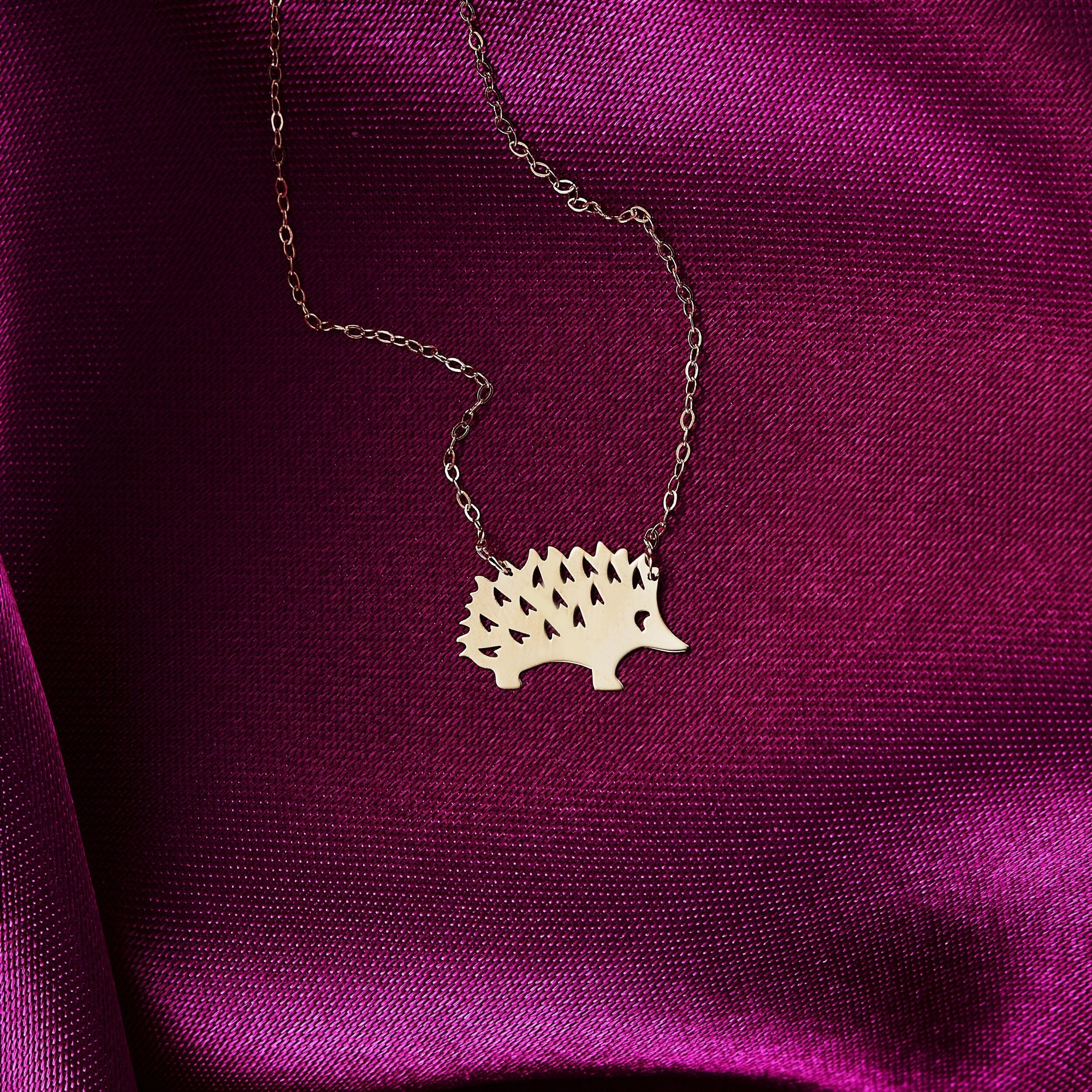 Close-up of a 14K gold hedgehog pendant necklace displayed on rich pink satin fabric.