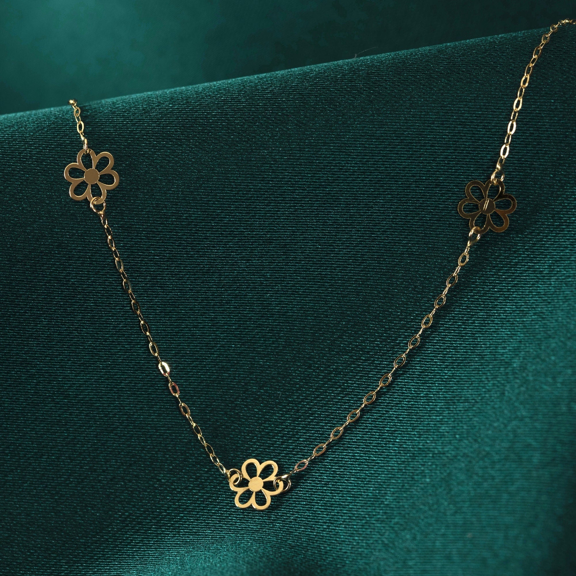 14K gold necklace featuring evenly spaced cut-out flower charms on a delicate chain, displayed on deep green satin.