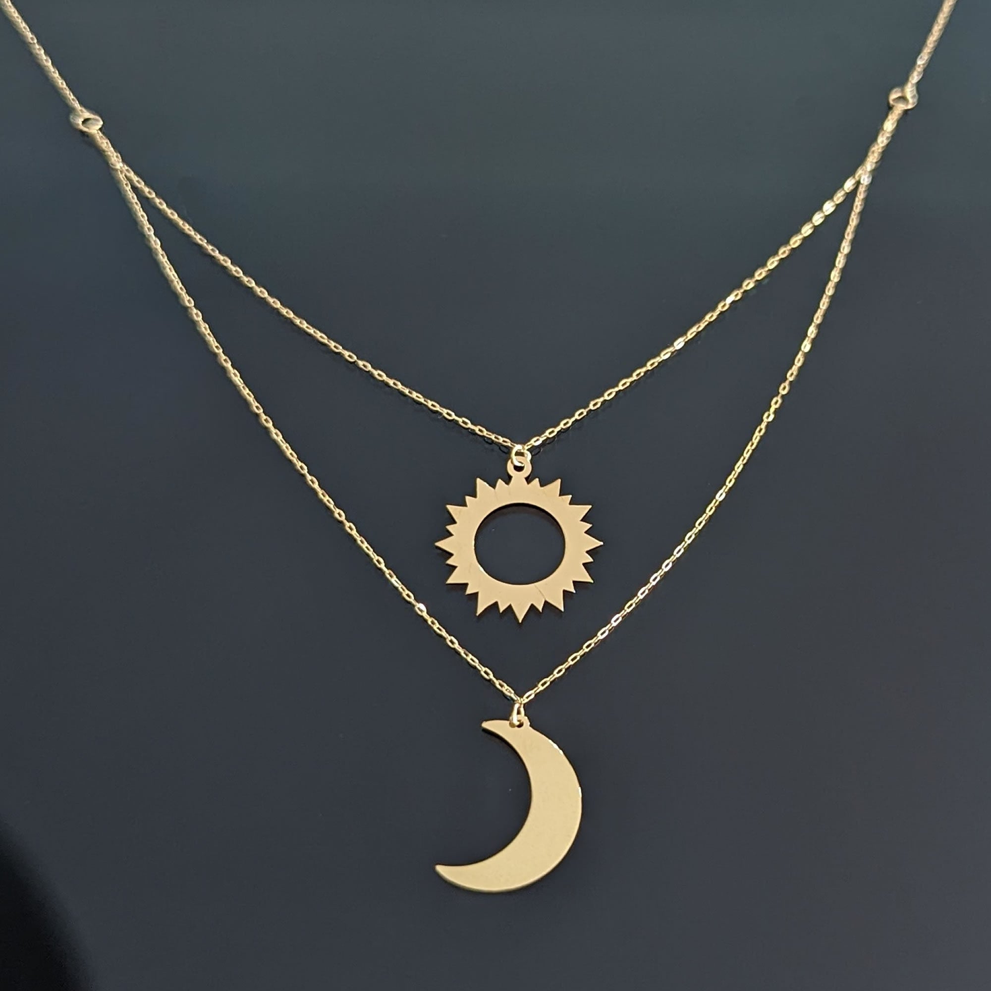 Layered 14K gold necklace with a polished crescent moon and cut-out sun pendant displayed on a dark reflective background.