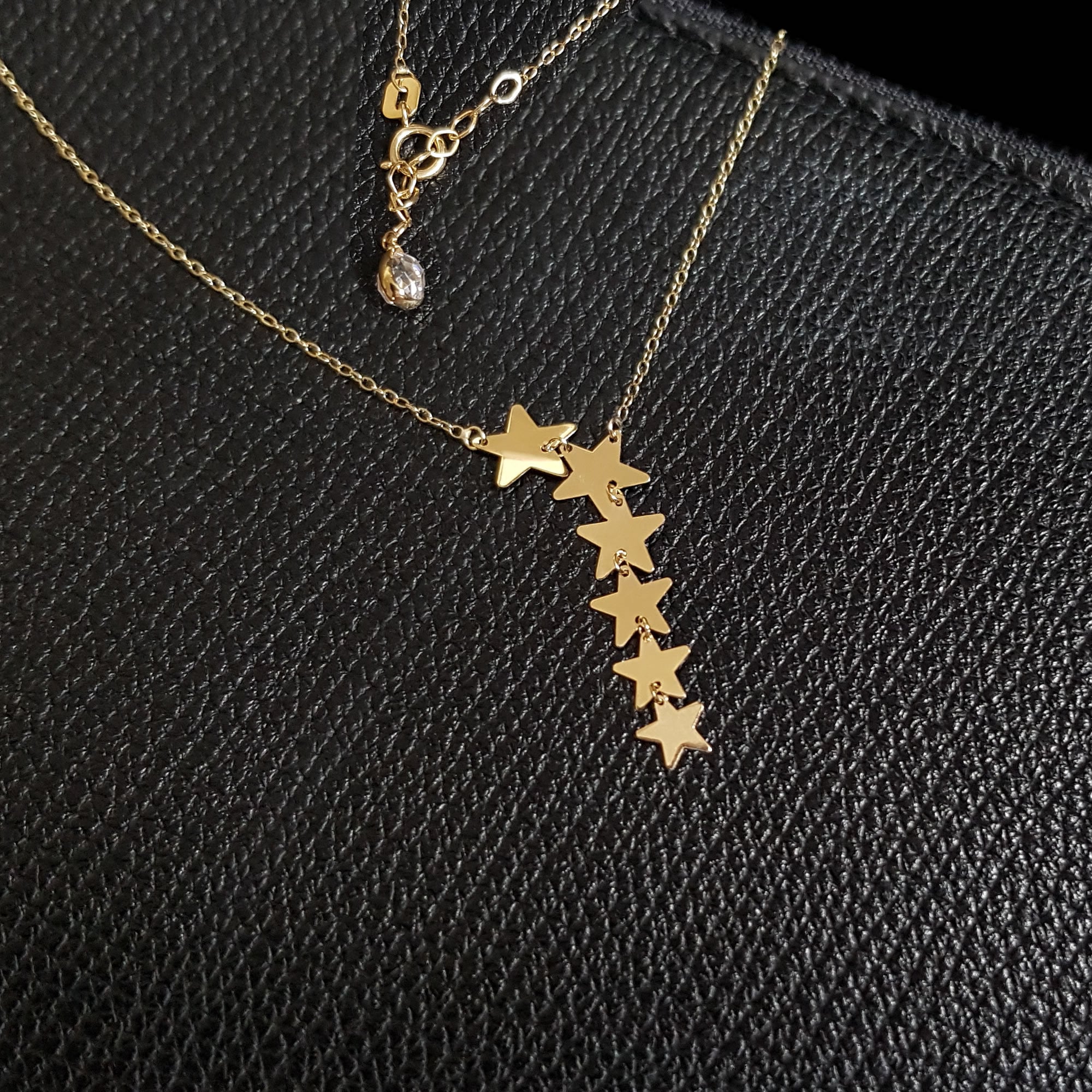 Seven-star cascade necklace in 14K gold displayed on black textured leather background, featuring minimalist celestial design.